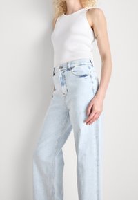 Woman with curly blonde hair wearing a white sleeveless ribbed top and light blue high-waisted wide-leg jeans against a plain background.