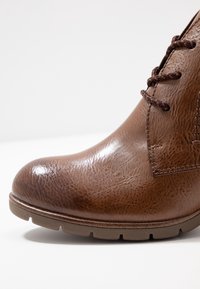 Brązowe skórzane botki za kostkę o teksturowanym wykończeniu, z okrągłym czubkiem i brązowymi sznurówkami. Posiadają kontrastującą gumową podeszwę z rowkami do przyczepności.