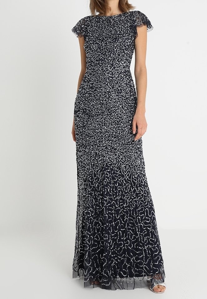 Tmavěmodré dlouhé šaty s stříbrnými korálkovými vzory, přiléhající v pase, s průsvitnými volnými rukávy, na stojící ženě.