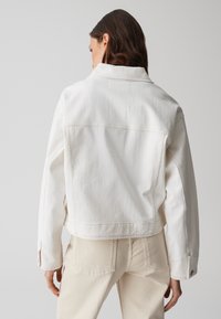 Giacca di denim bianco con colletto strutturato e cuciture visibili, dotata di due tasche laterali e un taglio leggermente accorciato.