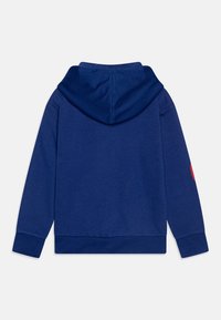 Vista posteriore di un felpa blu con cappuccio texturizzato e una toppa rossa sulla manica destra, adagiata su uno sfondo bianco.