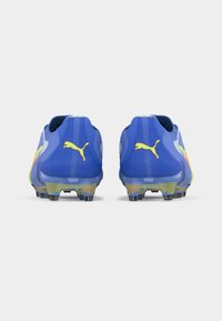 Ζευγάρι μπλε ποδοσφαιρικών παπουτσιών Puma, εμφανιζόμενα από πίσω, με κίτρινα λογότυπα Puma και πολύχρωμες εγκοπές σταταρίσματος σε λευκό φόντο.