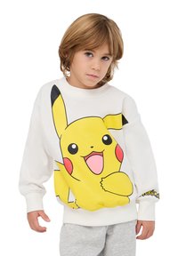 Felpa bianca con una grande grafica di Pikachu gialla sul davanti. Presenta polsini e orlo a costine, abbinata a pantaloni grigi.