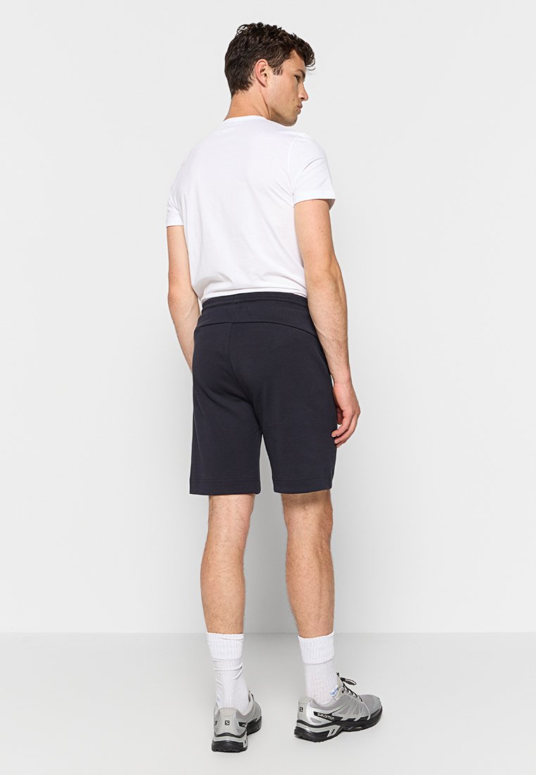 Shorts de sport bleu marine avec une taille élastique, coupés au niveau des genoux, associés à des chaussettes blanches et des baskets grises, vus de l'arrière.