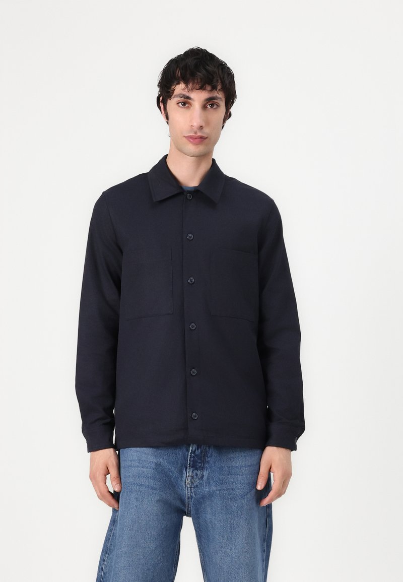 Marineblå skjorte med knapper, krave, to framlommer og lange ermer, laget av et glatt materiale. Kombinert med lyseblå jeans.