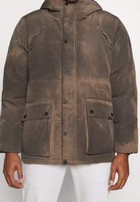 Brązowa pikowana kurtka z kapturem, wyposażona w czarne zatrzaski, dwie przednie kieszenie oraz miękkie, teksturowane wykończenie.