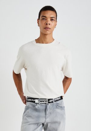 Balta medvilninė marškinėliai trumpomis rankovėmis, apvaliu kaklu, dėvimi su pilkais džinsais ir juodu diržu, ant kurio yra baltas tekstas.