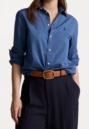 Chemise bleu à manches retroussées avec un petit logo. Associée à une ceinture marron et un pantalon foncé, mettant en avant un style décontracté.