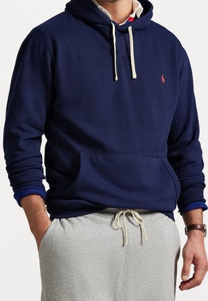Homme portant un sweat à capuche bleu marine avec un logo rose, des cordons blancs, un pantalon de survêtement gris, une chemise blanche à col et une montre-bracelet marron, mains dans les poches.