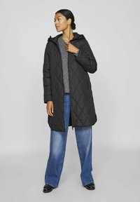 Svart vadderad lång kappa med huva, med ett diamantmönster, framzipper och sidofickor, bärs över en grå stickad tröja och blå jeans.