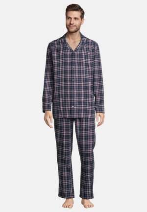 Mężczyzna stojący boso, ubrany w granatową, czerwoną i białą flanelową piżamę z długim rękawem zapinaną na guziki oraz dopasowane spodnie na białym tle.