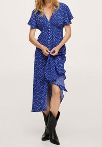 Sinivalkoinen pisteellinen mekko, jossa on lyhyet liehuvat hihat, napitus edessä, volangimainen helma ja reiden korkeudelle ulottuva halkio, yhdistettynä mustiin cowboy-kengiksiin.