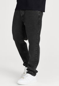Vaqueros de mezclilla negra con ajuste relajado, que presentan costuras visibles y piernas ajustadas, combinados con zapatillas deportivas blancas.