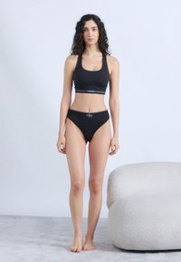 Γυναίκα με κατσαρά μαλλιά που φοράει μαύρο αθλητικό σουτιέν και εσώρουχο, όρθια ξυπόλητη δίπλα σε λευκή πολυθρόνα με υφή.