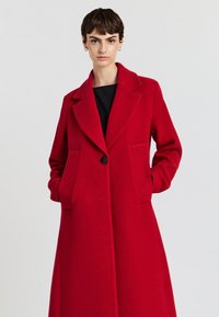 Roter Wollmantel mit langen Ärmeln, Reverskragen, vorderem Knopfverschluss und zwei vorderen Taschen. Glatte Textur und klassisches, mittellanges Design.