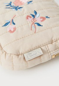 Quiltet stoffpose i beige, med rosa og blå blomsterbroderi og en liten etikett. Glidelås lukking langs bunnen.