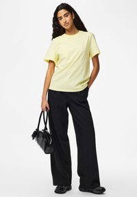 Jasnożółta bawełniana koszulka z krótkimi rękawami, zestawiona z czarnymi szerokimi spodniami w prążki. Modelka trzyma czarną torebkę.