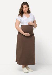 Zwangere jurk met een witte stippen-top en een lange bruine rok, voorzien van een V-halslijn en zachte, rekbare stof. Witte schoenen maken de look compleet.