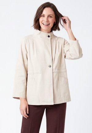 Donna sorridente che tocca i capelli, indossa una giacca beige chiaro con bottoni e pantaloni marrone scuro, in piedi davanti a uno sfondo bianco uniforme.