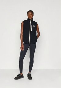 Zwarte fleecevest met hoge kraag en crèmekleurige ritssluiting, voorzien van een klein borstzakje. Gecombineerd met donkere leggings en zwarte sportschoenen.