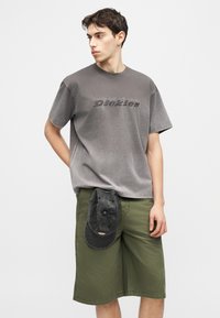 Νεαρός άνδρας που φοράει ένα ξεθωριασμένο γκρι μπλουζάκι Dickies και ελιά πράσινα σορτς με μαύρο καπέλο που έχει τοποθετηθεί στη ζώνη των σορτς.