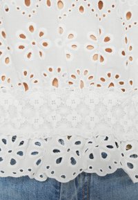 Tissu en coton blanc avec broderie œillet florale et bordure en dentelle, posé au-dessus d'un jean en denim bleu.