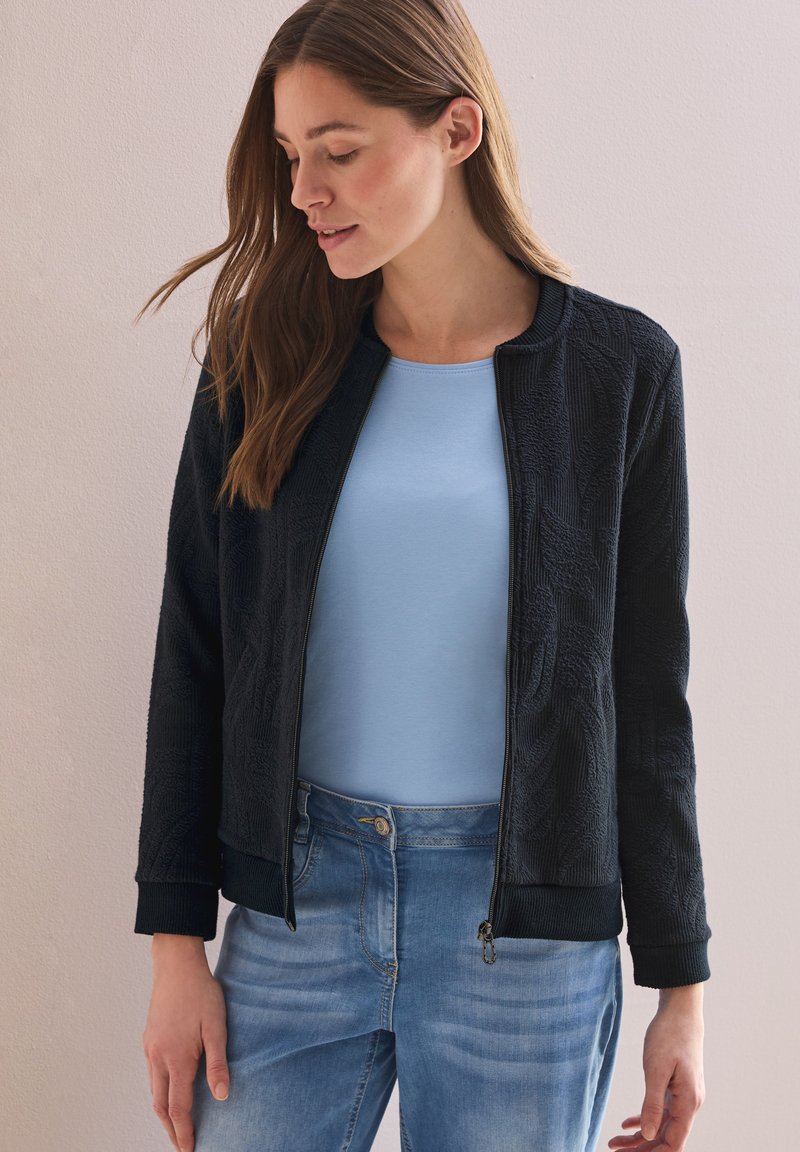 Chaqueta bomber texturizada negra con cierre frontal, puños acanalados y cuello redondo, llevada sobre una camiseta de color azul claro y pantalones de mezclilla azules.