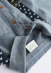 Blaue Jeanshemd mit leicht strukturiertem Material, ausgestattet mit braunen Knöpfen und einem marineblauen Riemen, der mit weißen Sterne verziert ist. Etiketten sind am Kragen sichtbar.