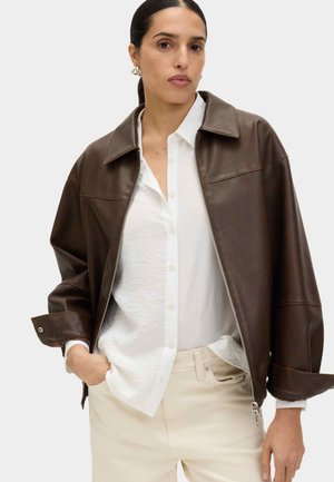 Vrouw met donker haar in een staart, draagend gouden oorringen in de vorm van ringen, een bruine leren jas met rits over een wit overhemd met knopen, en beige broek.