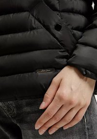Czarna kurtka puchowa z pikowanym wzorem, błyszczącym wykończeniem i małą metalową metką marki, z ręką opartą na tkaninie.
