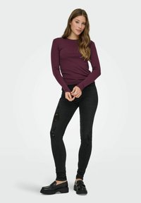 Bordeauxrotes Langarmoberteil, schwarze ripped Skinny-Jeans und schwarze Slipper. Das Oberteil hat einen figurbetonten Schnitt; die Jeans weisen auffällige Waschungen auf.