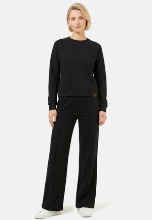 Kobieta stojąca przodem, ubrana w czarną bluzę z długim rękawem, pasujące szerokie spodnie i białe trampki na jednolitym tle.