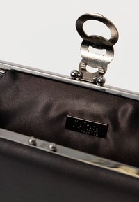 Τσάντα clutch με μεταλλικό πλαίσιο, που διαθέτει εσωτερικό από λείο, σκούρο ύφασμα. Το πλαίσιο έχει μια διακριτή κούμπωμα και μικρά διακοσμητικά στοιχεία.