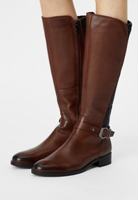 Brown leather knee-high boots with a side zipper, ribbed fabric accents, and a decorative metal buckle on the ankle strap. Smooth texture.