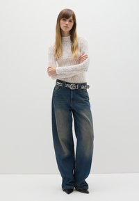 Top lungo bianco in pizzo con maniche lunghe abbinato a jeans blu in denim larghi. La cintura presenta accenti metallici decorativi. La modella è in piedi con le braccia incrociate.