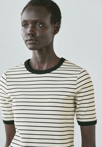 Junge schwarze Person mit kurzen lockigen Haaren, die ein weißes T-Shirt mit dünnen schwarzen horizontalen Streifen und schwarzem Rand am Kragen und an den Ärmeln trägt.