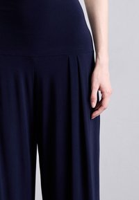 Navyblå gulvlængde kjole med en tætsiddende overdel og bred, flydende nederdel med plisseringer. Glat, elastisk stof. Hånd hvilende på nederdelen.