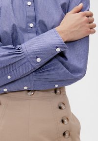 Sininen raidallinen napillinen paita, jossa on valkoiset napit, yhdistettynä beigeihin korkeavyötäröisiin housuihin, joissa on suuret koristenaapit sivulla.