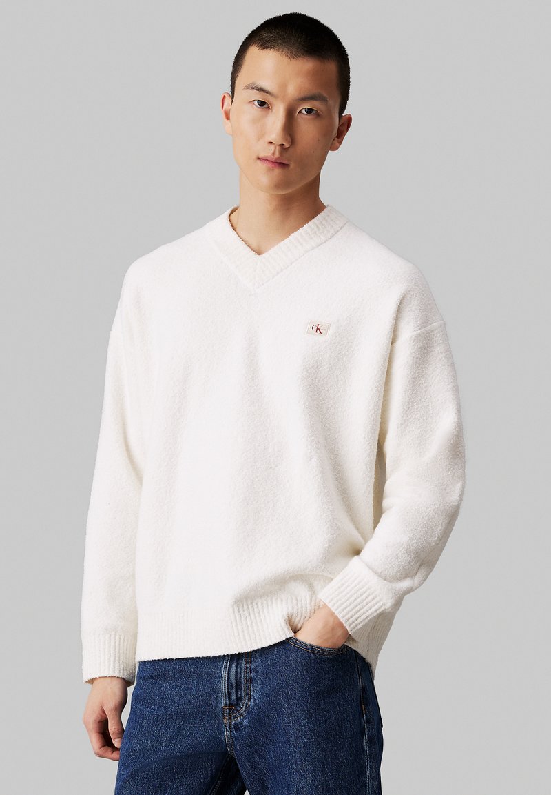 White v-neck sweater with a textured, fuzzy surface and ribbed cuffs, featuring a small logo patch on the chest. Worn with blue jeans.