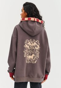 Hnedá voľná mikina s kapucňou a grafickým potlačou znázorňujúcou mytologické tvory a text "Expecto Patronum." Kapucňa má červený a béžový károvaný vzor.