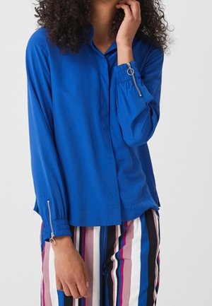 Kvinde iført en klar blå bluse med lange ærmer med lynlåse ved manchetterne og flerfarvede bukser med lodrette striber, der står mod en ensfarvet baggrund.