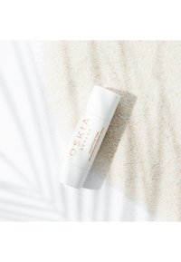 Oskia Adaptive Tan, en vit cylindrisk tube med guldtext, placerad på ljus beige sand, som framhäver en jämn struktur och ren design.