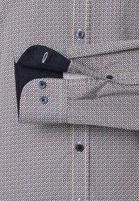 Hemd mit Knopfverschluss, das ein geometrisches Muster in Marineblau, Weiß und Rostfarben aufweist. Beinhaltet eine kontrastierende dunkle Tasche und schwarze Knöpfe.