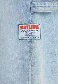 Tissu en denim bleu clair avec un patch rectangulaire blanc affichant "BITUME" en rouge et "solid" en gris en dessous, encadré par un motif à carreaux.