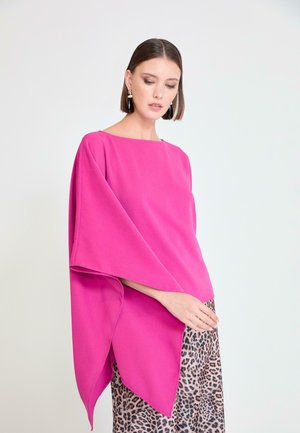 Mujer con poncho asimétrico de color rosa brillante y mangas anchas sobre falda de estampado de leopardo, mirando hacia abajo, fondo claro.