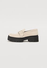 Beige Wildleder Slipper mit einer abgerundeten Spitze, ausgestattet mit einer massiven schwarzen Gummisohle und einem dekorativen Riemen mit einem silbernen Schnallenakzent.