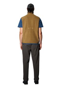 Bruine fleece vest met borduursel op de rug, gedragen over een blauwe T-shirt, gecombineerd met donkere broek. Eenvoudig ontwerp, geen zichtbare hardware of zakken.