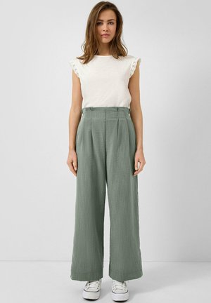 Femme portant un haut sans manches blanc avec bordure en dentelle, un pantalon vert taille haute à jambes larges, et des baskets blanches, debout devant un fond uni.