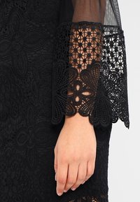 Robe noire avec des détails en dentelle complexe et une manche en tulle à motifs. La manche se termine par un ourlet en dentelle à bords festonnés.