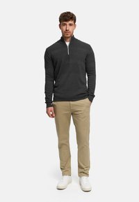 Mand iført en mørkegrå tekstureret sweater med kvart lynlås, beige bukser og hvide sneakers stående foran en ensfarvet hvid baggrund.